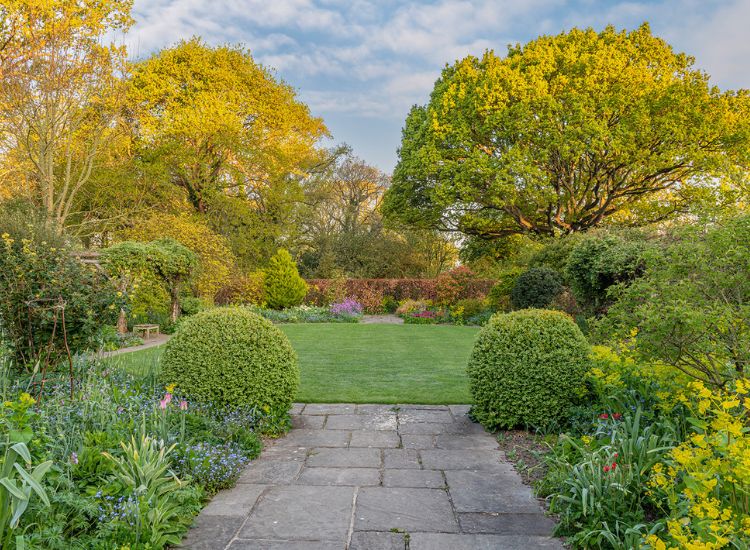 Stone path leading through a manicured garden with neatly trimmed shrubs, colourful flower borders, and mature trees in soft sunlight.